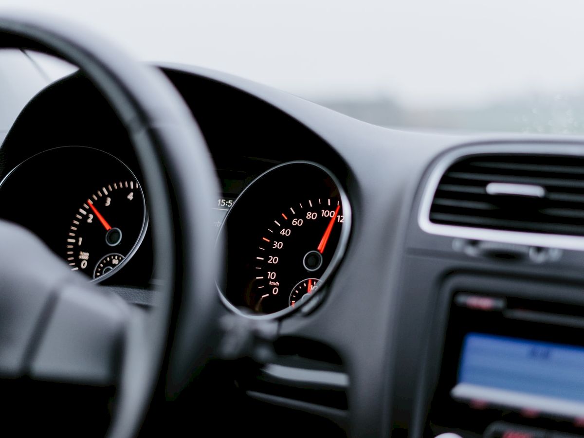 The image shows a car dashboard including the speedometer, tachometer, and an infotainment screen next to the steering wheel, displaying driving information.