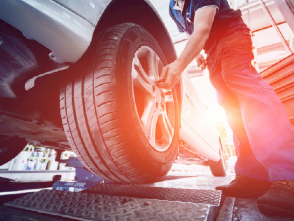 A mechanic is working on a tire of a car that is lifted, with sunlight streaming in from the background inside a well-lit workshop.