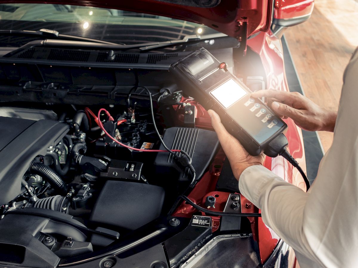 The image shows a person using a diagnostic tool to check the engine of a car with its hood open.