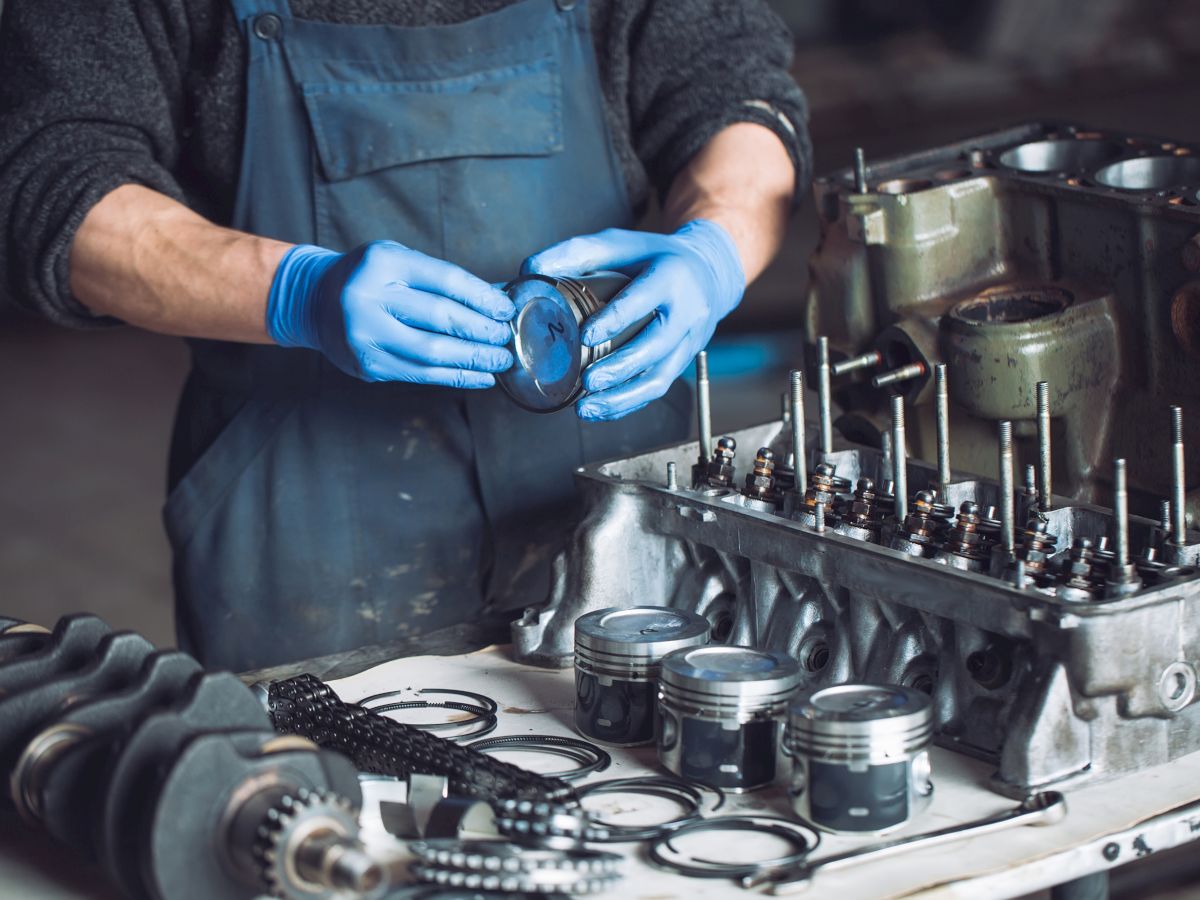 A person wearing blue gloves and an apron is working on engine parts, including pistons and a crankshaft, at a workbench indoors.