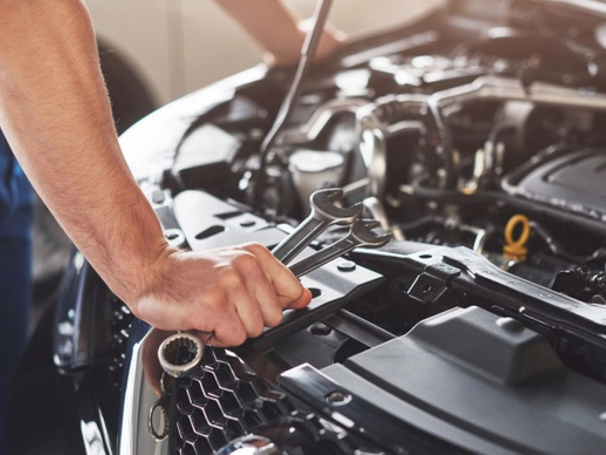 A mechanic is working on a car engine, holding a wrench. The image shows the engine compartment and the man's arm using the tool.