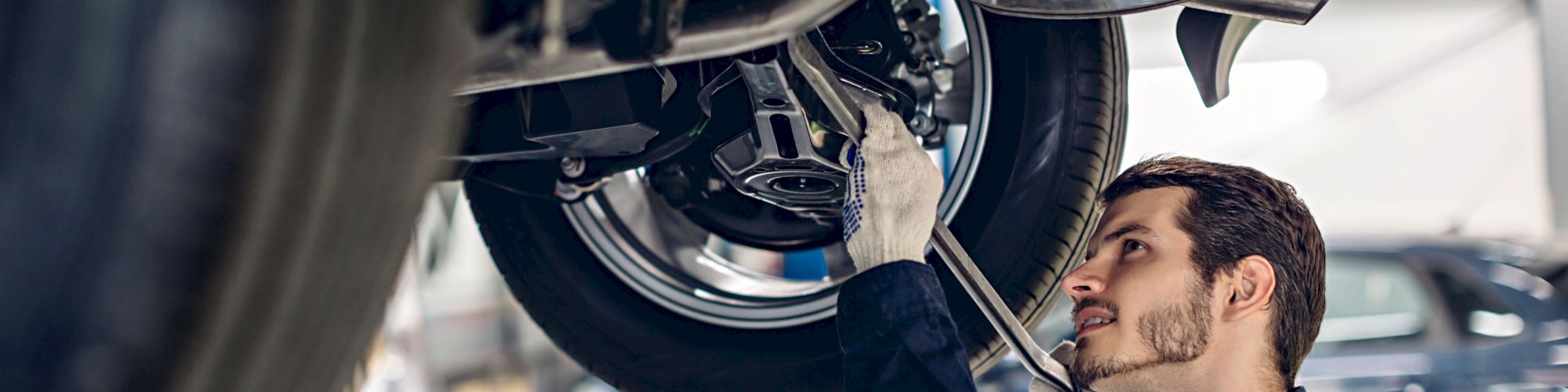 A mechanic is working underneath a car held up by a hydraulic lift in a garage, using a wrench to adjust parts near the wheel.