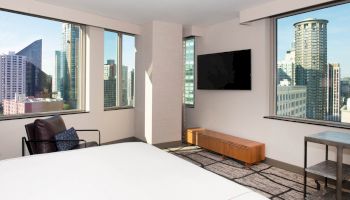 A modern hotel room with a bed, armchair, wall-mounted TV, and large windows offering a cityscape view of tall buildings.