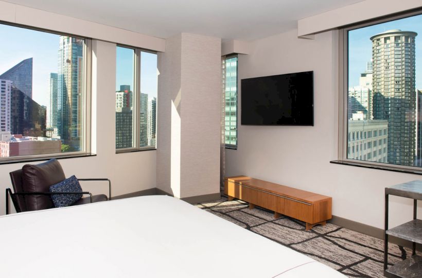 A modern hotel room with a bed, armchair, wall-mounted TV, and large windows offering a cityscape view of tall buildings.