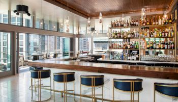 A stylish, modern bar with elegant seating, a well-stocked selection of bottles, and large windows offering a view of the cityscape ends the sentence.