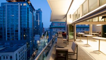 The image shows a modern urban balcony overlooking a city street with tall buildings. The balcony has chairs and tables with city lights visible.