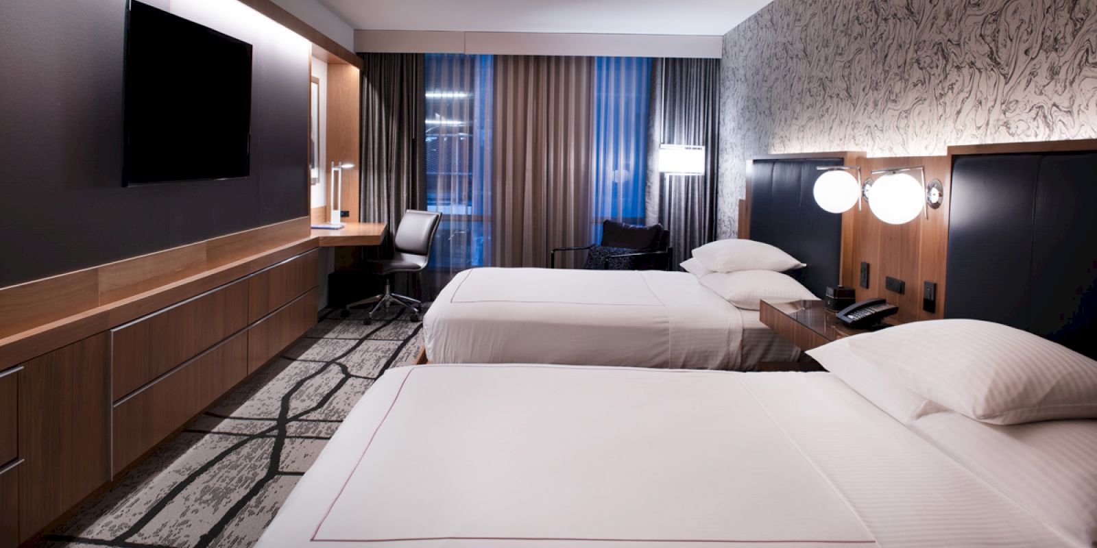 The image shows a modern hotel room with two neatly made beds, a wall-mounted TV, a work desk with a chair, and stylish decor.