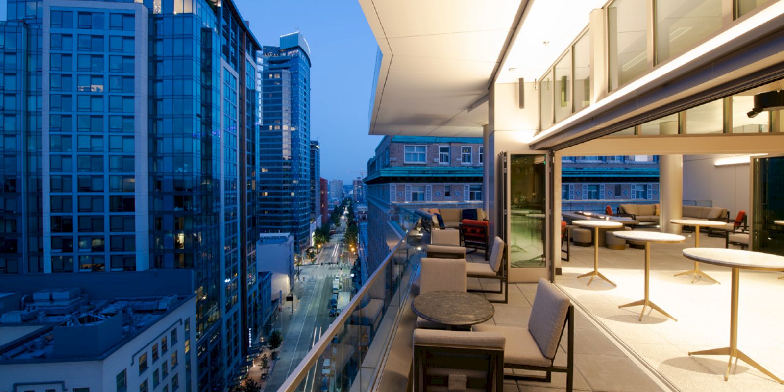The image shows a modern cityscape from a balcony with seating and dining areas, overlooking buildings and a street during twilight.