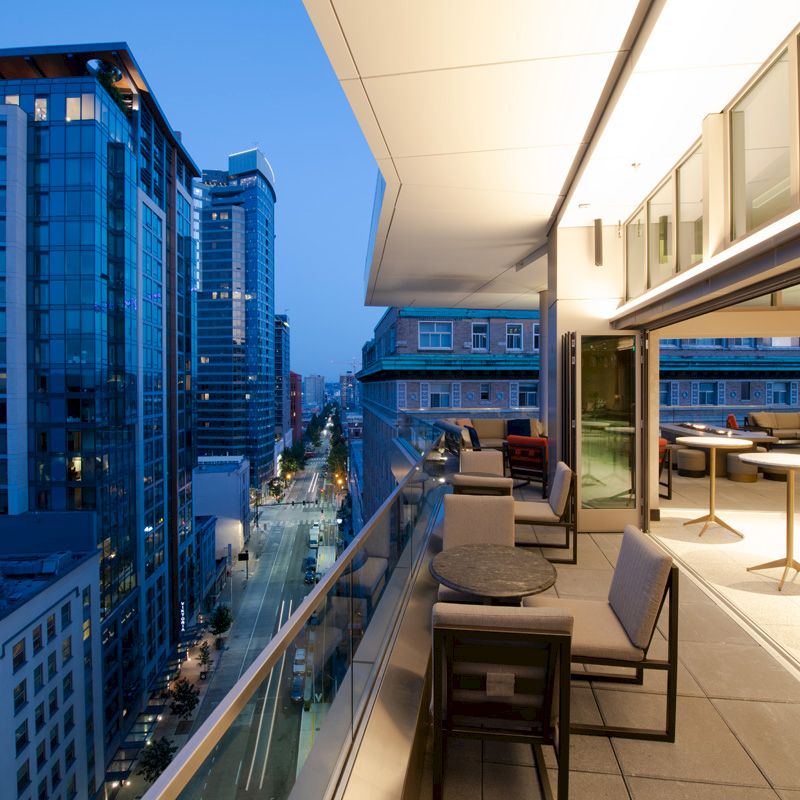 A modern cityscape during dusk with high-rise buildings and a balcony featuring tables and chairs overlooking the street below.