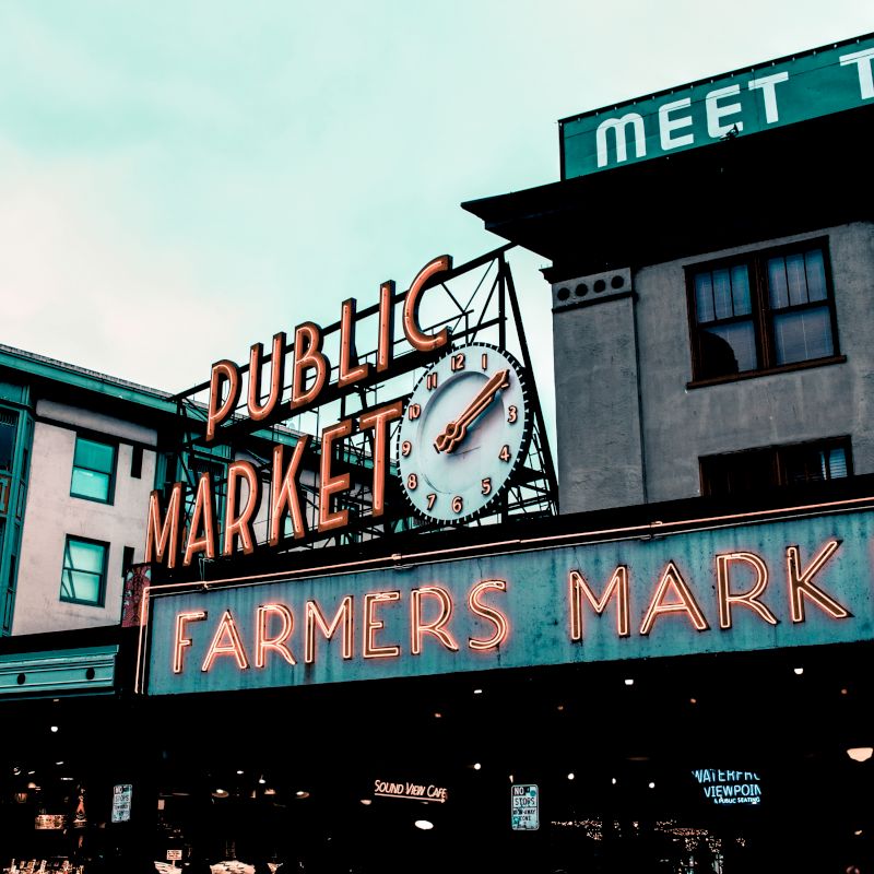 The image shows a public market sign and a farmers market sign, along with a clock, on an urban street, with buildings in the background.
