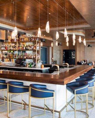 The image shows a modern bar with a wooden counter, blue barstools, shelves with bottles, hanging lights, and a lounge area in the background.