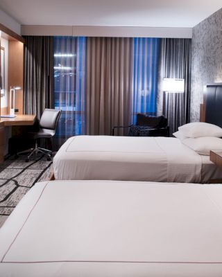 A modern hotel room with two beds, a TV, a desk with a chair, lamps, and large windows with brown curtains, ending the sentence.