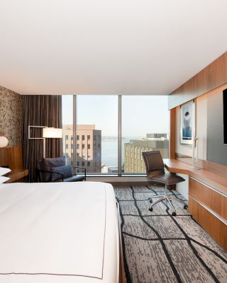 This image shows a modern hotel room featuring a large bed, a desk with a chair, a flat-screen TV, and a city view through floor-to-ceiling windows.