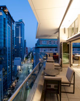 A modern cityscape with tall buildings is viewed from a balcony at dusk. The balcony features outdoor seating and tables, softly lit.