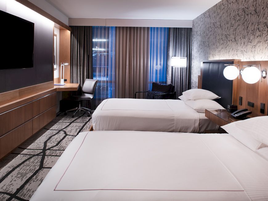 A modern hotel room with two beds, a TV, a desk with a chair, and lamps on the nightstands, featuring a patterned wall and floor.