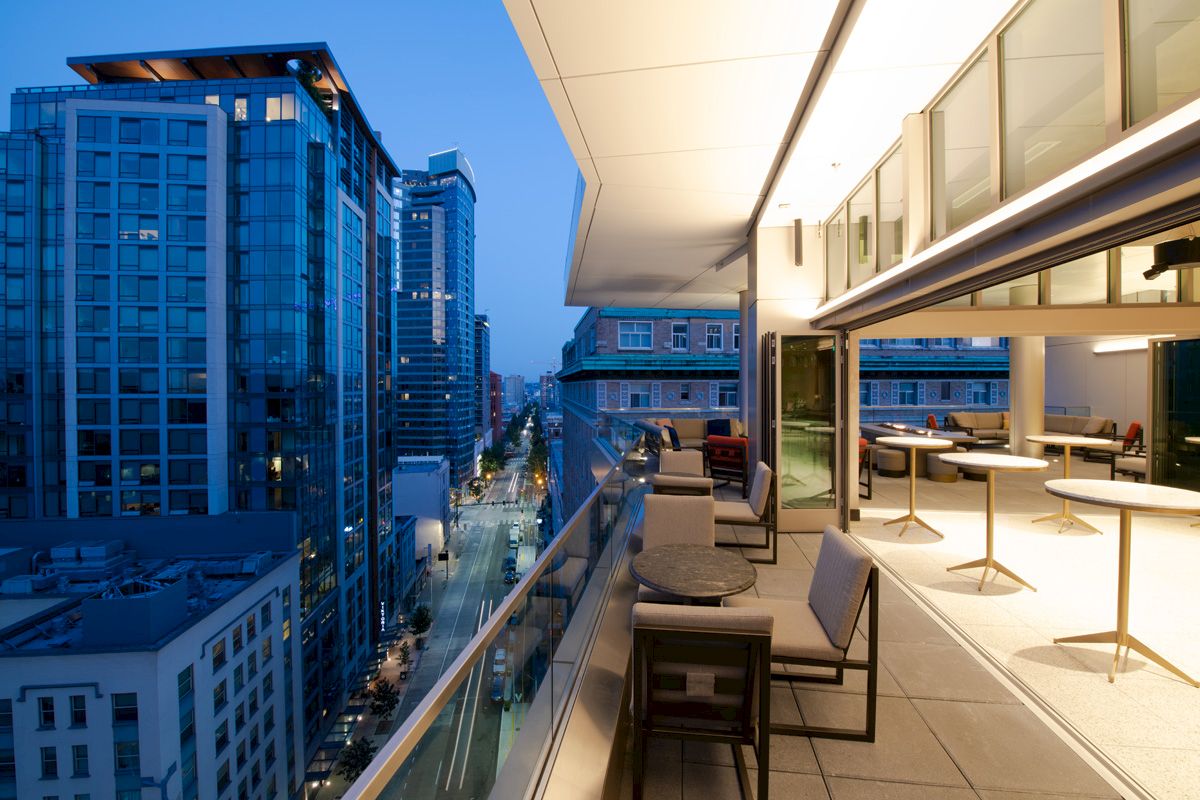 A modern cityscape with tall buildings is viewed from a balcony with tables, chairs, and floor-to-ceiling windows at dusk.
