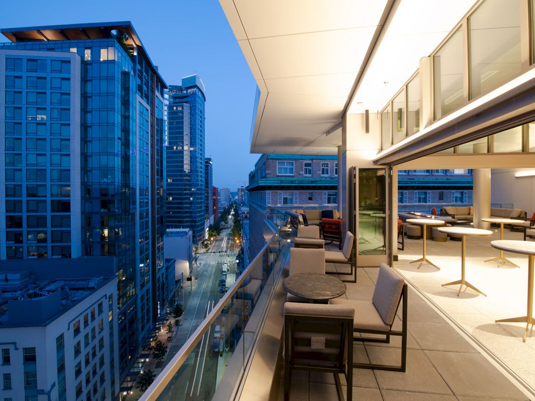 A modern cityscape with tall buildings is viewed from a balcony with tables, chairs, and floor-to-ceiling windows at dusk.