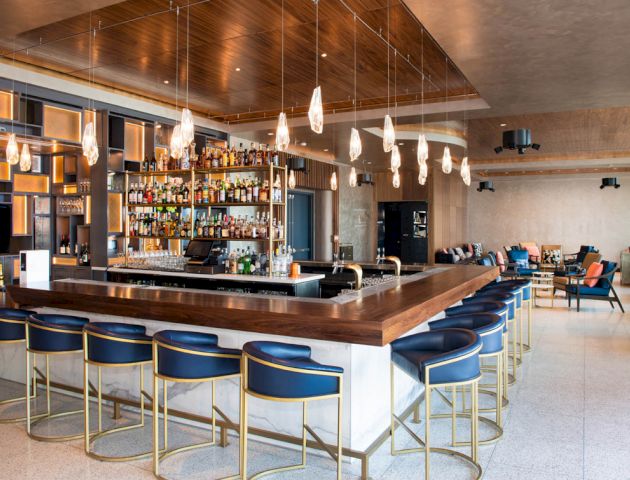 This image features a modern bar with a stylish wooden counter and blue barstools, surrounded by hanging lights and a variety of bottles on shelves behind.