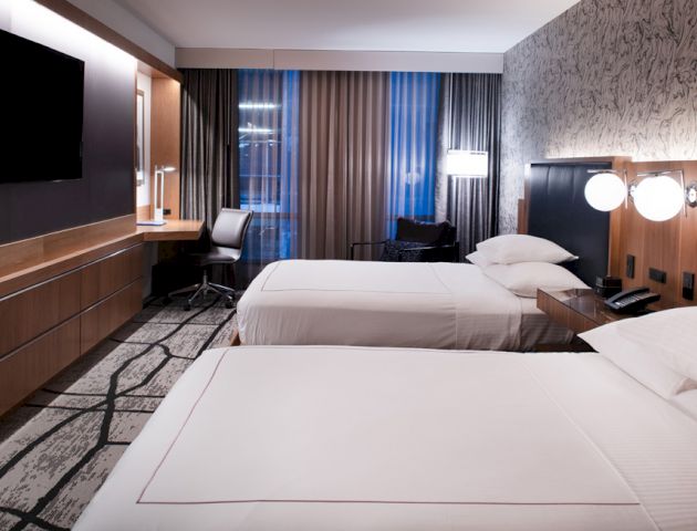 A modern hotel room with two neatly made beds, a wall-mounted TV, a desk with a chair, large windows with curtains, and contemporary decor.