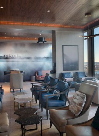 A modern lounge with numerous chairs and small tables facing large windows that offer a view of a bay and a Ferris wheel outside.