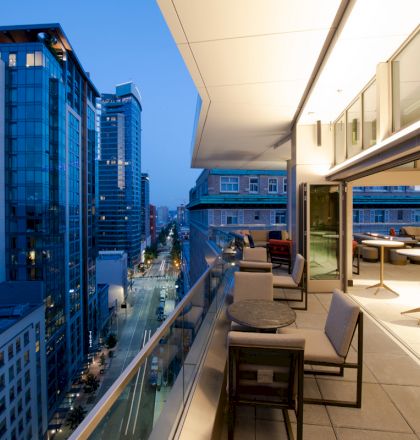 The image shows a modern outdoor terrace of a high-rise building with seating areas, overlooking a cityscape at dusk.