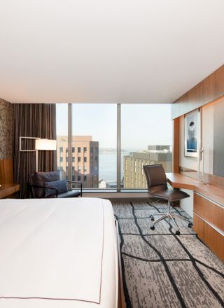 A modern hotel room with a large bed, desk, chair, flat-screen TV, and a city view through a large window.