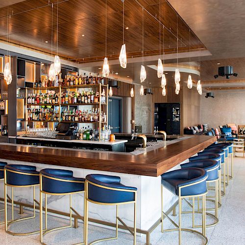 The image shows a modern, stylish bar with a wooden counter, blue cushioned bar stools, and hanging lights, surrounded by various seating areas.