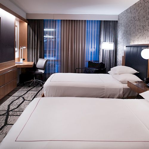 A modern hotel room with two beds, a large TV, a desk with a chair, and patterned wallpaper.