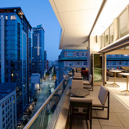 A modern rooftop balcony with seating offers views of an urban cityscape filled with high-rise buildings as evening approaches.