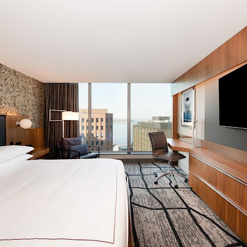 A modern hotel room with a large bed, wall-mounted TV, desk, and chair. The room features floor-to-ceiling windows with a city view.