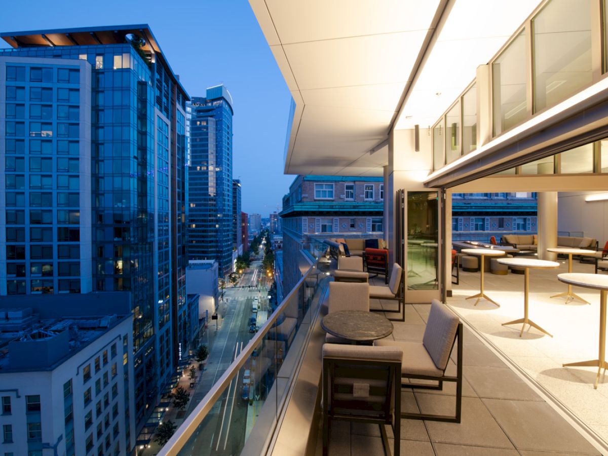 This image shows a modern balcony at dusk with a cityscape view, featuring outdoor seating and tables, and adjacent buildings lit up.