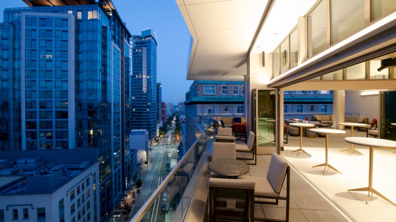 This image shows a modern balcony at dusk with a cityscape view, featuring outdoor seating and tables, and adjacent buildings lit up.