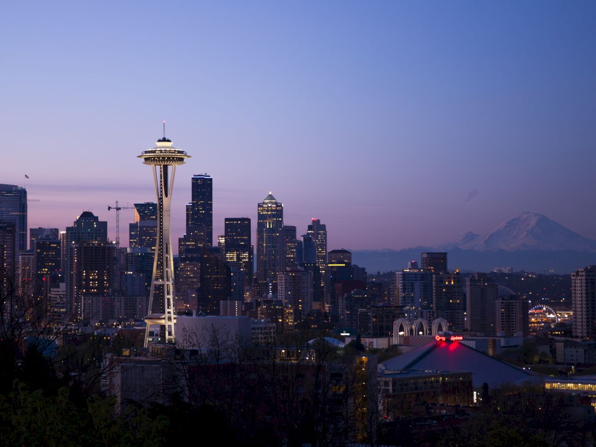 This image depicts the Seattle skyline at dusk, featuring the illuminated Space Needle and downtown buildings with Mount Rainier visible in the background.