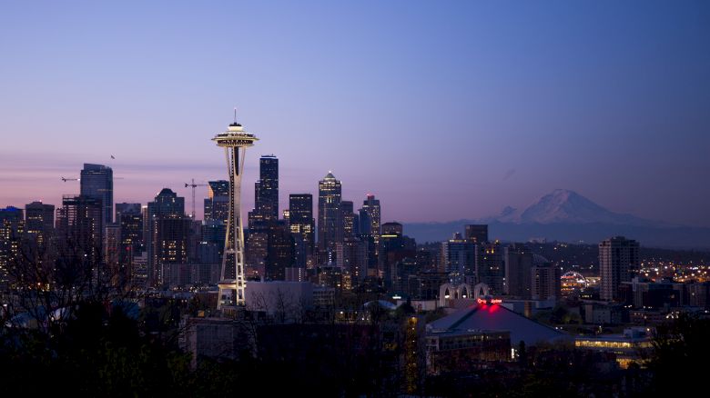 This image depicts the Seattle skyline at dusk, featuring the illuminated Space Needle and downtown buildings with Mount Rainier visible in the background.