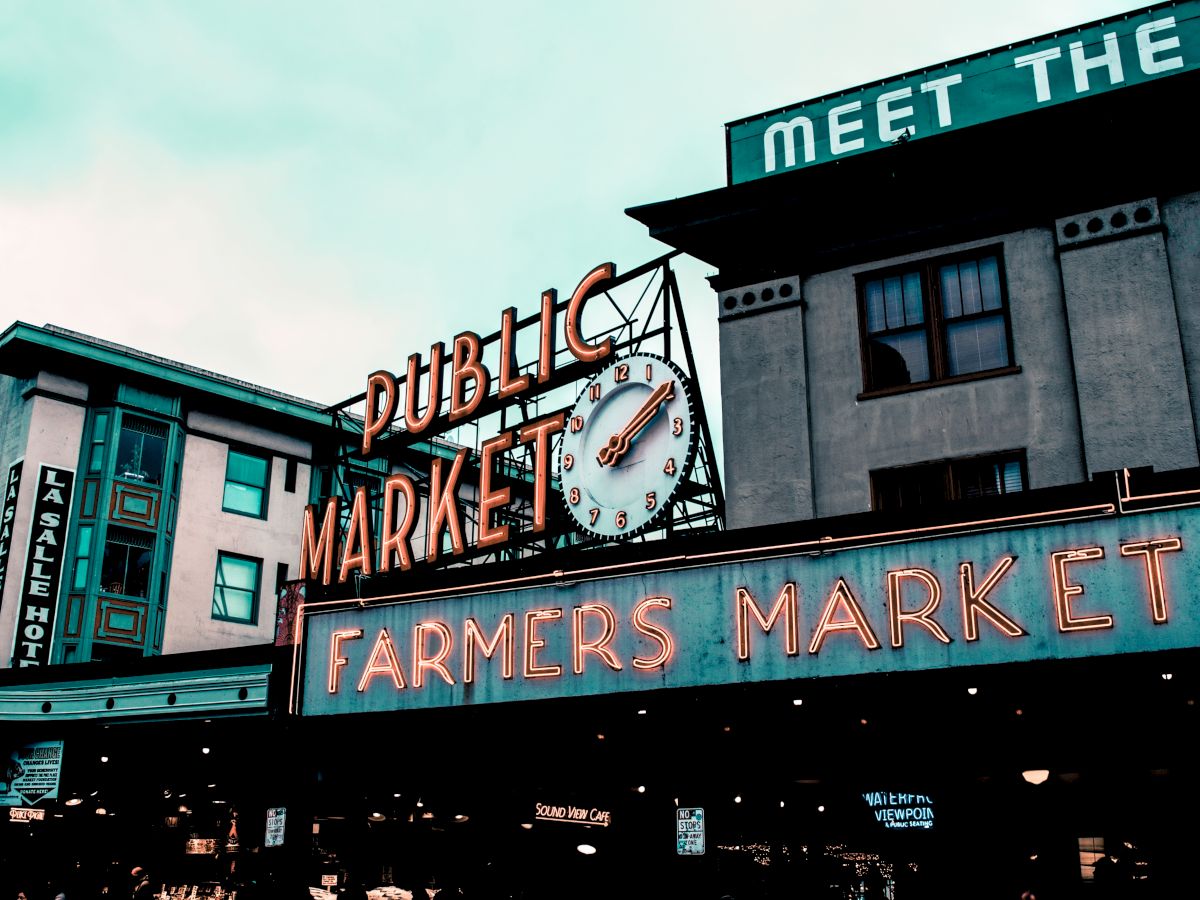 The image depicts the neon sign of a public farmers market with a clock. The surrounding buildings contribute to a vibrant, urban atmosphere.