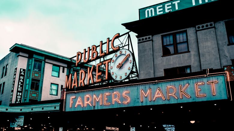 The image depicts the neon sign of a public farmers market with a clock. The surrounding buildings contribute to a vibrant, urban atmosphere.