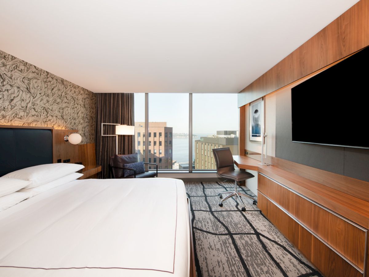 A modern hotel room with a large bed, wall-mounted TV, desk, chair, and window with a cityscape view. Room features wood and neutral tones.