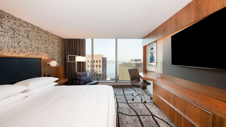 A modern hotel room with a large bed, wall-mounted TV, desk, chair, and window with a cityscape view. Room features wood and neutral tones.