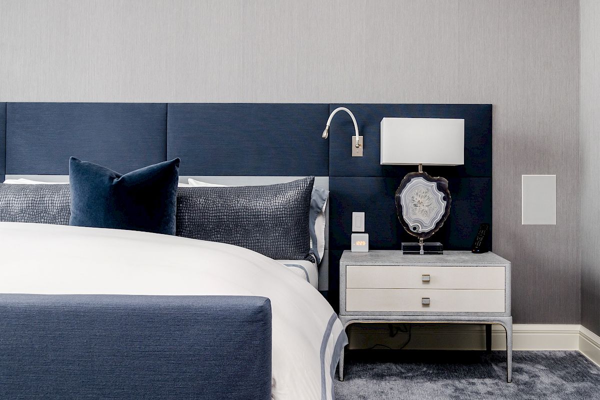 A modern bedroom with a navy blue and white color scheme, featuring a bed with a padded headboard and a nightstand with a lamp and decor.