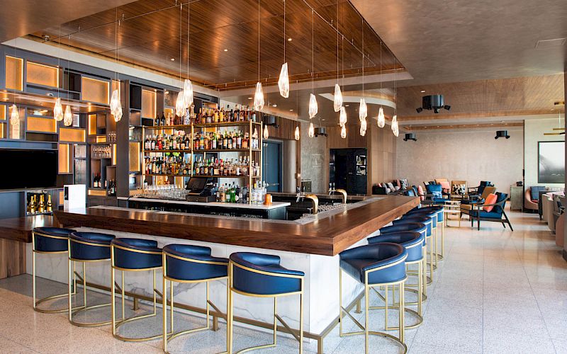 A modern bar with a wooden counter, blue chairs, hanging lights, and a well-stocked liquor shelf in a spacious, elegantly designed setting.