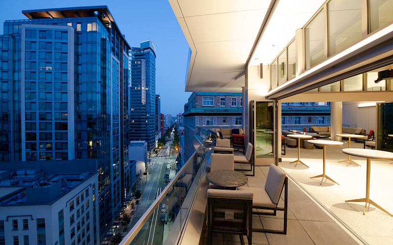 A modern balcony overlooking a cityscape at dusk, with seating areas and tables, adjacent to an illuminated indoor space through large windows.