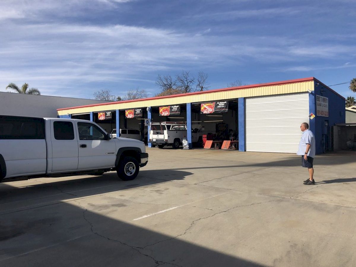 A man stands in a parking lot with a white pickup truck outside a building with open garage bays showcasing vehicles inside.