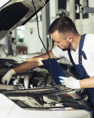 A mechanic in a workshop is inspecting the engine of a white car with the hood open, using a blue tool and wearing gloves and overalls.