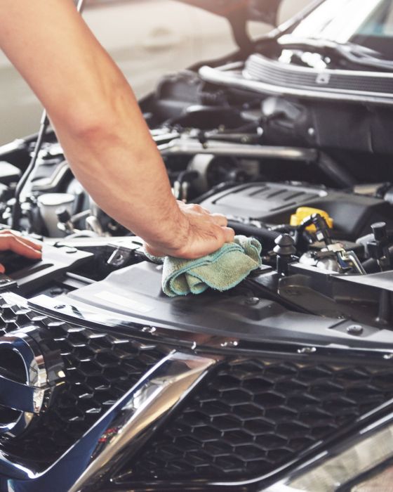 A person is cleaning the inside of a car's engine bay with a cloth. The car's hood is open, revealing various engine components.