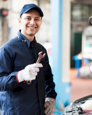 A mechanic in blue overalls and a cap smiles while holding a wrench, standing next to an open car hood in a workshop.