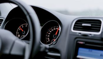 The image shows the dashboard of a car, including the steering wheel, speedometer, tachometer, and part of the center console with a screen.