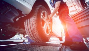 A mechanic is working on a car, adjusting a tire in a garage with bright sunlight in the background.