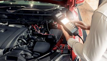 A person is using a diagnostic tool to check the engine of a red car with its hood open, indicating an inspection or maintenance activity.