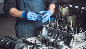 A person wearing blue gloves is assembling or inspecting engine parts on a workbench, including pistons, rings, and engine components.