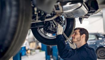 A mechanic in a garage is working under a vehicle, using a wrench to adjust or fix parts while looking up intently.